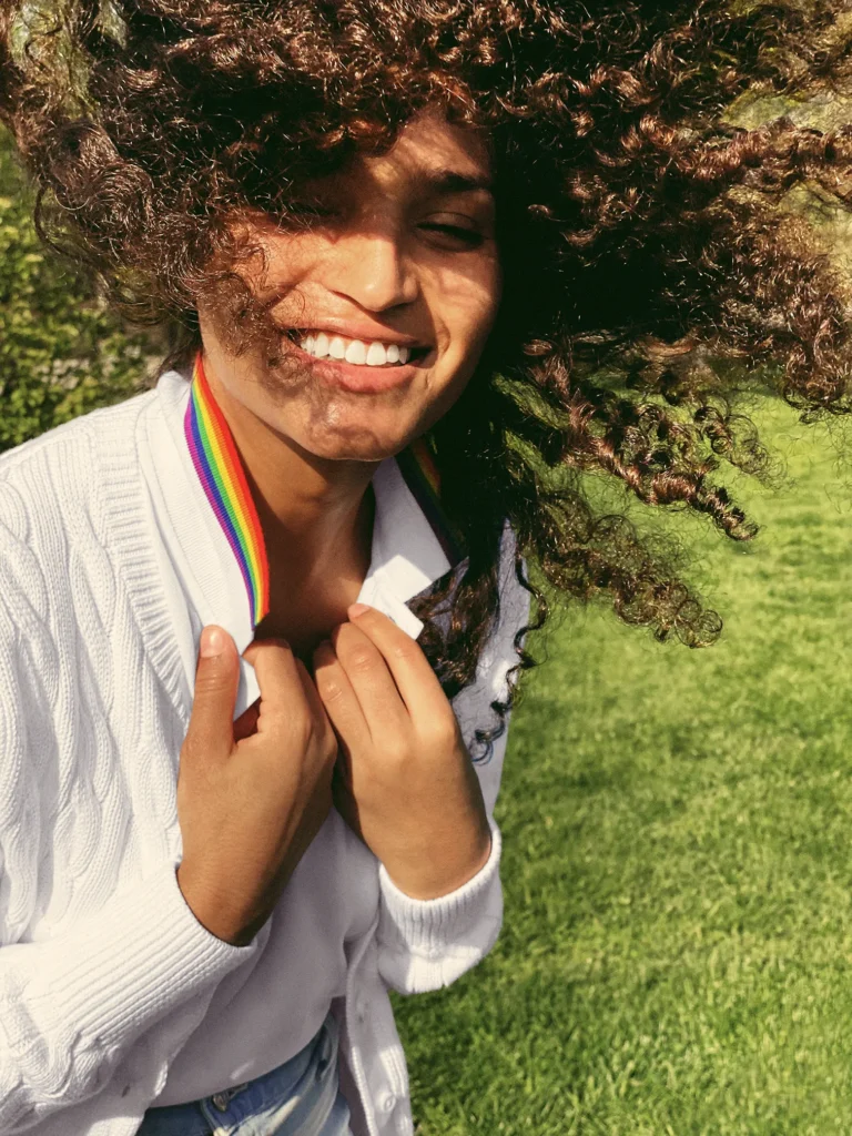 Indya Moore is captured in a candid moment outdoors, smiling with windblown curly hair. They wear a white cable-knit cardigan over a white polo shirt, with a rainbow-striped collar peeking out. Sunlight casts a glow on their face, highlighting their joyful expression.