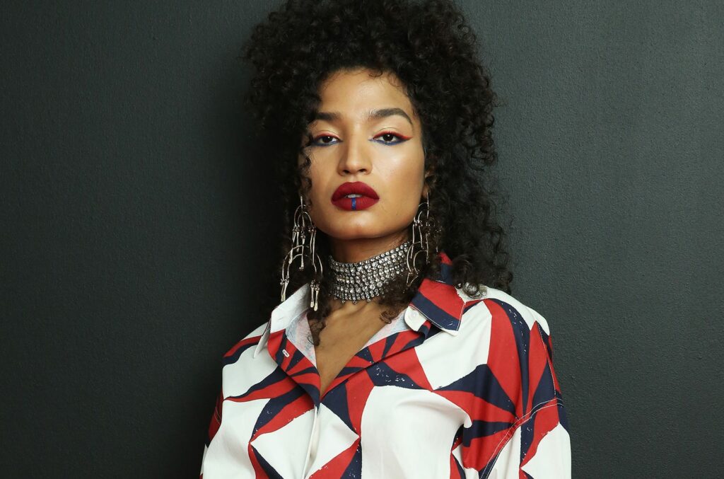 Indya Moore poses against a dark background, showcasing bold makeup with red and blue eyeliner, deep red lipstick featuring a blue accent on the lower lip, and sharply defined eyebrows. They wear large, intricate metallic earrings and a sparkling rhinestone choker. Their outfit is a button-up shirt with a striking red, white, and navy geometric pattern. Their expression is confident and intense, with a direct gaze at the camera.