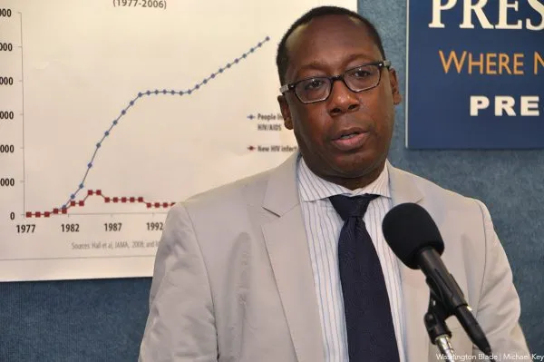 Cornelius Baker speaks at a press conference, standing in front of a chart displaying data on HIV/AIDS cases. He wears glasses, a light-colored suit, and a dark tie while addressing an audience with a serious expression.