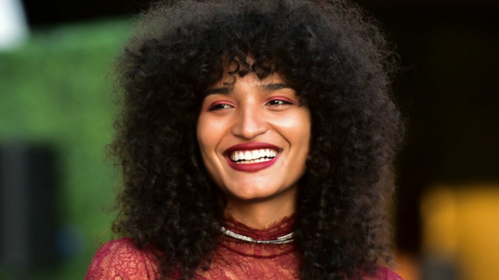 Indya Moore smiles radiantly while wearing a red lace high-neck top. Their voluminous curly hair frames their face, and they have bold pink eyeshadow and deep berry lipstick. The blurred green background suggests an outdoor event, adding warmth and vibrance to the image.