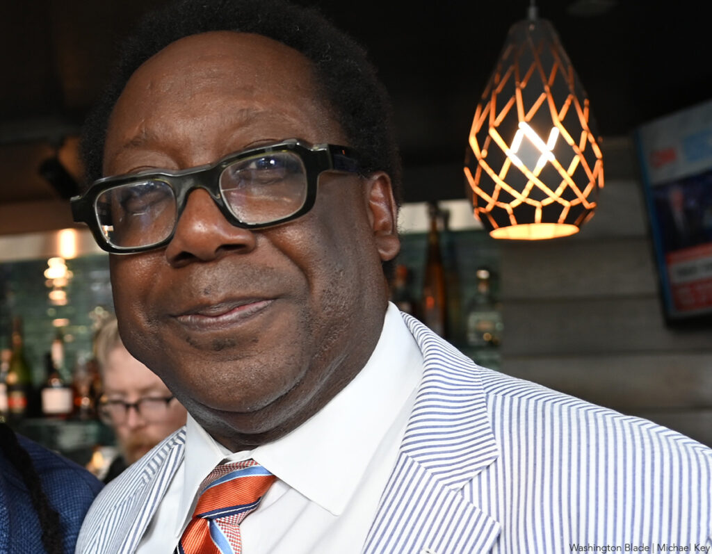 Cornelius Baker is seen at a social event, wearing a white and blue striped suit with a bright orange tie. He has a relaxed expression with a slight smile, and the background features dim lighting and a hanging lamp.