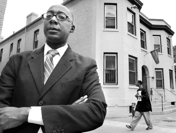 A black and white portrait of Cornelius Baker standing with arms crossed in front of a brick building with a pride flag. He wears a suit and glasses, looking confidently at the camera. In the background, a person walks past the building.