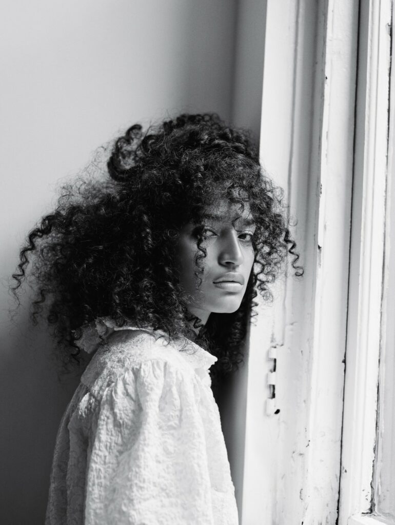 A black and white photograph of Indya Moore standing by a window, gazing into the distance with an introspective expression. Their curly hair cascades naturally, partially obscuring their face. They wear a textured white blouse with a high collar and ruffled details. Soft natural light from the window highlights the contours of their face, creating a moody and artistic atmosphere.