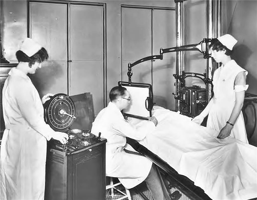 A black-and-white historical photograph showing Dr. Alan L. Hart seated at an X-ray machine while two nurses assist. One nurse monitors a control panel while the other stands beside a patient lying under a white sheet on an examination table. The large, mechanical X-ray apparatus surrounds the patient. The scene reflects early 20th-century medical technology and Hart's pioneering work in radiology.