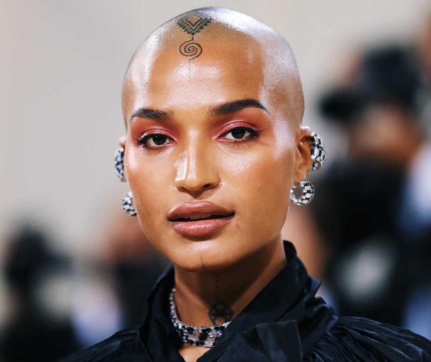  Indya Moore looks poised at a red carpet event, wearing a black high-collared outfit with sparkling silver earrings. Their shaved head features a delicate geometric tattoo at the center of their forehead. Their makeup includes soft, warm eyeshadow and glossy lips, creating an elegant and futuristic look.