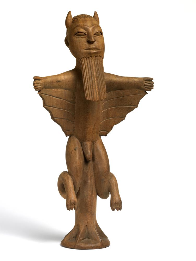 A carved wooden figurine of a Sasabonsam, a vampiric creature from West African folklore. The figure has a humanoid form with bat-like wings extending from its arms, small horns on its head, and a long, vertical beard. It is mounted on a tree-like base, symbolizing its association with forest-dwelling spirits. The stylized carving gives it an ancient and ritualistic appearance.