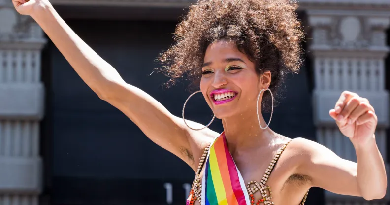 Indya Moore joyfully raises their arms while participating in World Pride 2019 in New York. They wear a studded gold dress with a rainbow sash and large hoop earrings. Their curly hair is styled voluminously, and they have bright pink lipstick and shimmering eye makeup. The background features classic urban architecture.