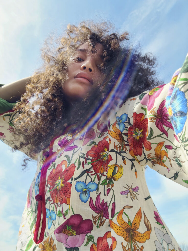 Indya Moore takes a striking low-angle selfie against a bright blue sky, wearing a flowing cream-colored outfit adorned with colorful floral and insect illustrations. Their curly hair is slightly windblown, and a lens flare creates a rainbow arc across the image. Their gaze is calm and introspective.