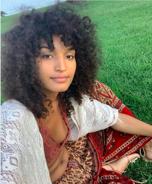 Indya Moore takes a relaxed outdoor selfie while sitting on vibrant green grass. They wear a bohemian-inspired outfit, including a red patterned skirt, a lace bralette, and a white crochet cardigan draped over their shoulders. Their curly hair cascades naturally, and they have a serene, makeup-free expression.