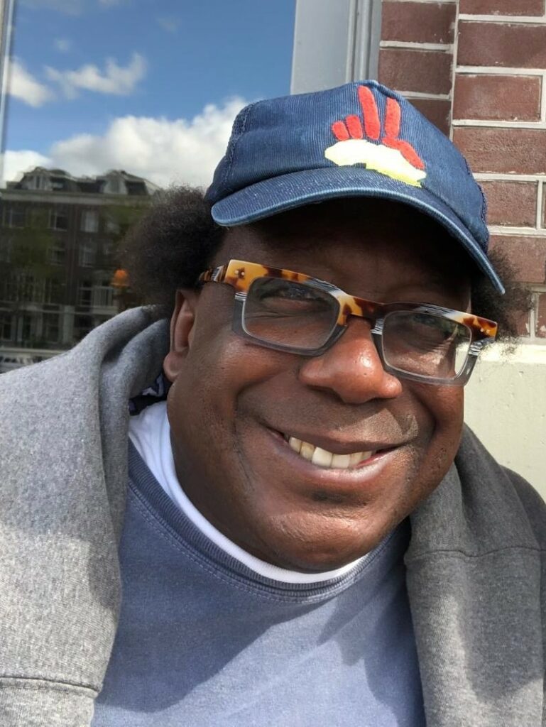 Cornelius Baker smiles warmly while wearing a navy blue baseball cap with a peace symbol, tortoiseshell glasses, and a gray sweatshirt. The background includes a cityscape with windows reflecting the sky.