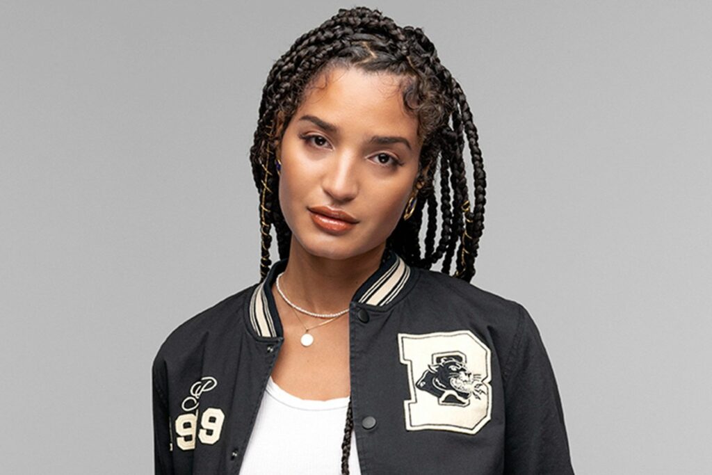 Indya Moore poses in a black varsity jacket with a bold embroidered patch, worn over a white top. Their hair is styled in long, tight braids with gold accents. They wear minimal yet glowing makeup, with a natural lip color and soft eyeliner. Their expression is calm and confident against a simple gray backdrop.