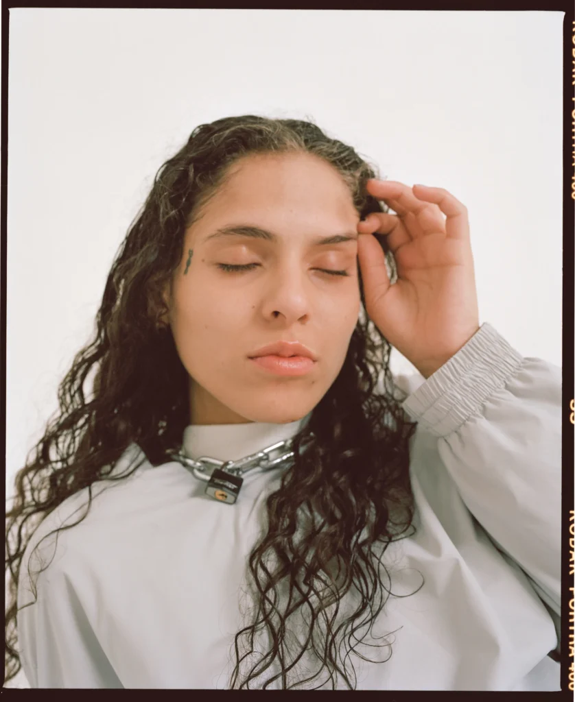070 Shake poses with her eyes closed, her hand gently touching her forehead. She wears a silver chain necklace with a small padlock and a light-colored oversized jacket. Her long, wavy hair cascades over her shoulders against a plain white background.