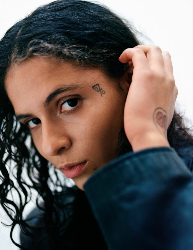 A close-up portrait of 070 Shake with wavy hair as she gazes into the camera. A small "love" symbol tattoo is visible near her temple. She wears a dark jacket, and her hand slightly tugs at her hair.