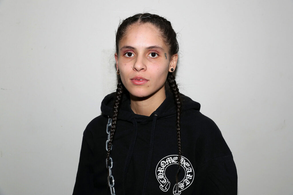 070 Shake poses in a black hoodie with "Chrome Hearts" printed on the front. Her hair is styled in two long braids, and she has subtle eye makeup with a slightly serious expression. The background is a plain off-white wall.
