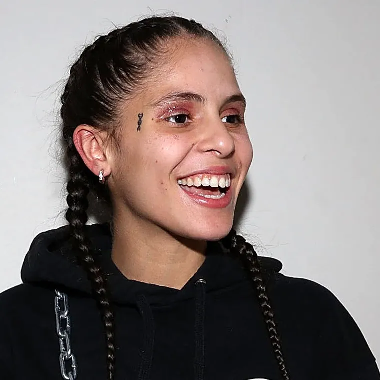 070 Shake smiles brightly, showing her teeth, while wearing a black hoodie with metallic chain details. Her hair is styled in two braids, and a small tattoo is visible near her eye.