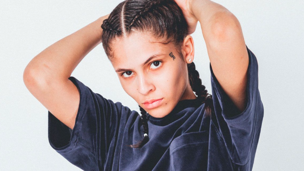 070 Shake looks intensely into the camera while pulling her hair back with both hands. She wears a dark velvet top, and her face tattoo is prominently visible.
