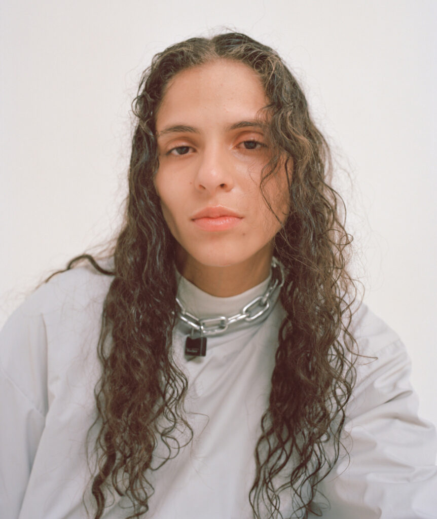 A soft-lit portrait of 070 Shake wearing a silver padlock necklace and a light-colored oversized jacket. Her expression is calm, and her long, wavy hair frames her face.