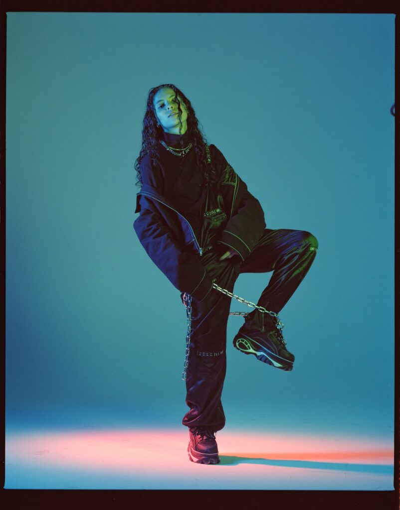 070 Shake poses dynamically in a studio with blue and green lighting, wearing an oversized black outfit with metallic chains. She lifts one knee and tilts her head slightly, with her long, wavy hair framing her face.