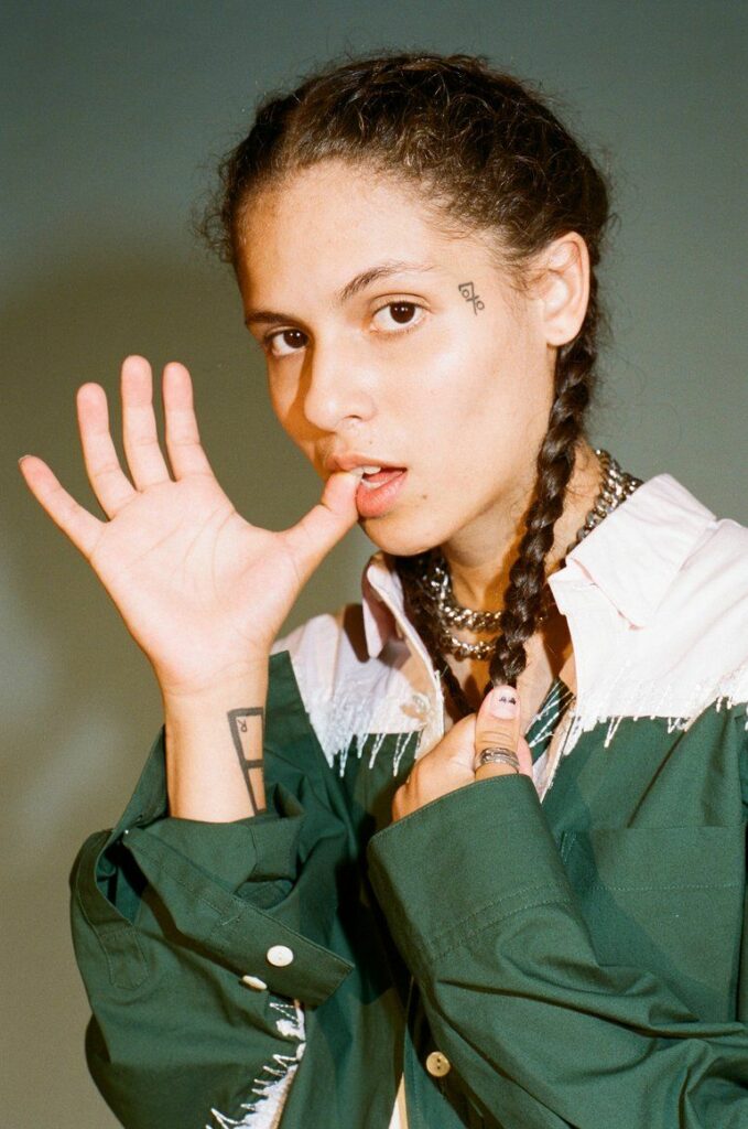 070 Shake playfully bites her thumb while wearing a green and white button-up shirt. Her hair is styled in two braids, and multiple tattoos are visible on her arms and face. The lighting casts a warm glow on her face.