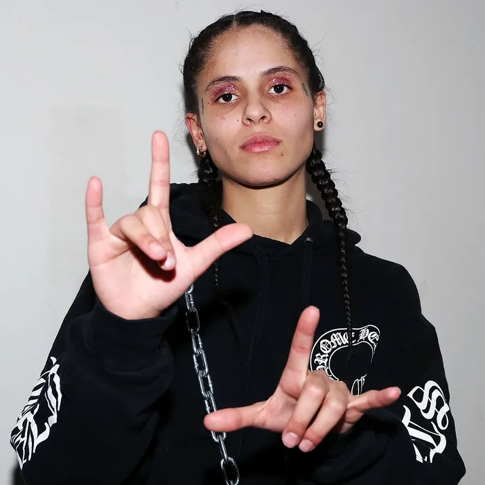 070 Shake wears a black "Chrome Hearts" hoodie and raises her hands in a gesture, forming shapes with her fingers. She maintains a calm and confident expression while looking directly at the camera. The background is a simple, off-white wall.