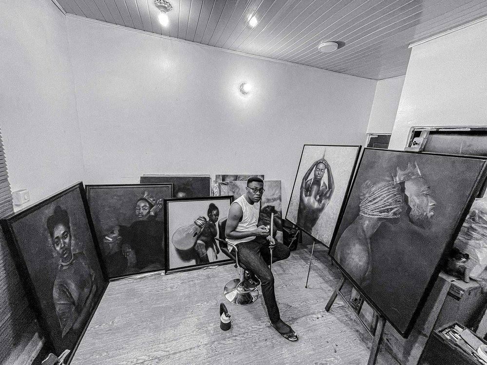 A black-and-white image of artist Alex Peter Idoko sitting on a stool in his studio, surrounded by multiple large-scale pyrography artworks leaning against the walls. His pieces depict highly detailed portraits, showcasing his mastery of fire-based art techniques.