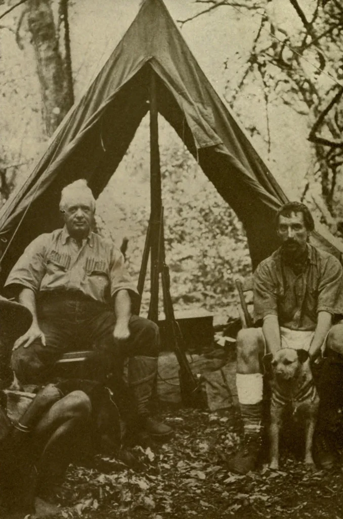 A historical photograph of Edgar Beecher Bronson (1856–1917) and John Alfred Jordan (1857–1933) at a hunting camp. The two men are seated outside a tent with rifles, a dog, and a local guide, symbolizing their exploration and colonial-era hunting expeditions.