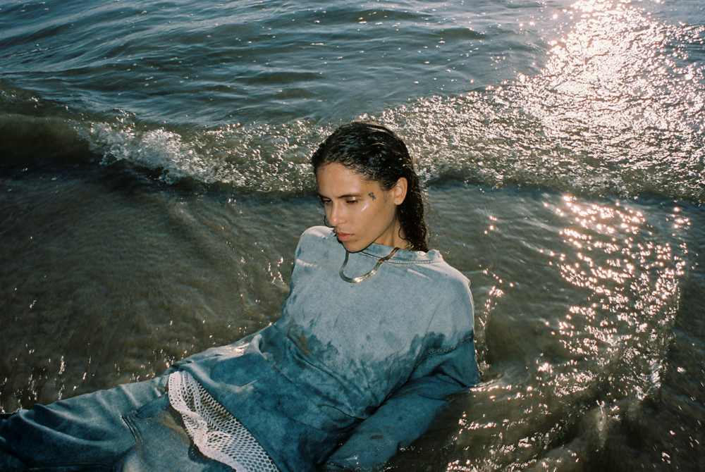 070 Shake sits partially submerged in water, wearing a faded blue oversized sweater and mesh details. Her wet hair clings to her face as she gazes downward, with the sunlight creating a sparkling effect on the water’s surface.
