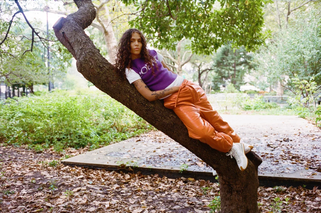 070 Shake lounges against a slanted tree in a park, wearing a purple knitted vest over a white T-shirt and bright orange pants. Her curly hair cascades over her shoulder as she gazes directly at the camera in a relaxed pose.