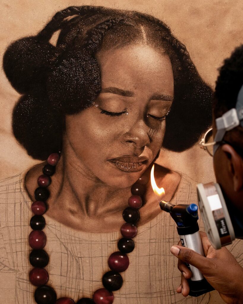 The artist Alex Peter Idoko is seen using a small torch to burn intricate details into a pyrography artwork of a woman with traditional African hair braids and large beaded jewelry. The image captures his unique artistic process and precision.