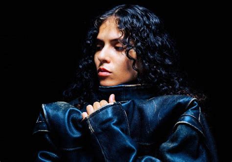 A close-up portrait of 070 Shake wearing a heavy oversized dark jacket, her hands pulling the collar up. Her curly hair frames her face as she looks off to the side with an intense expression against a black background.