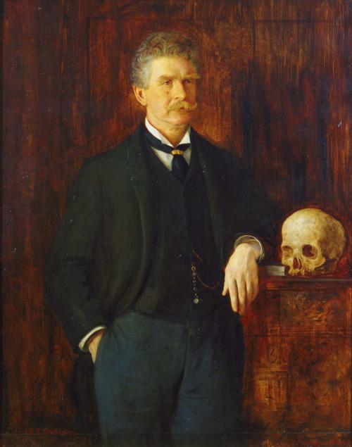 A formal oil painting of Ambrose Bierce standing beside a table with a human skull. He wears a dark three-piece suit with a pocket watch chain and rests one hand on the table. The background is wooden and richly textured.