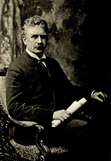 A seated black and white photograph of Ambrose Bierce holding a rolled document in one hand. He sits in an ornate chair and wears a dark suit with a high-collared white shirt and tie. The backdrop is a textured studio background.