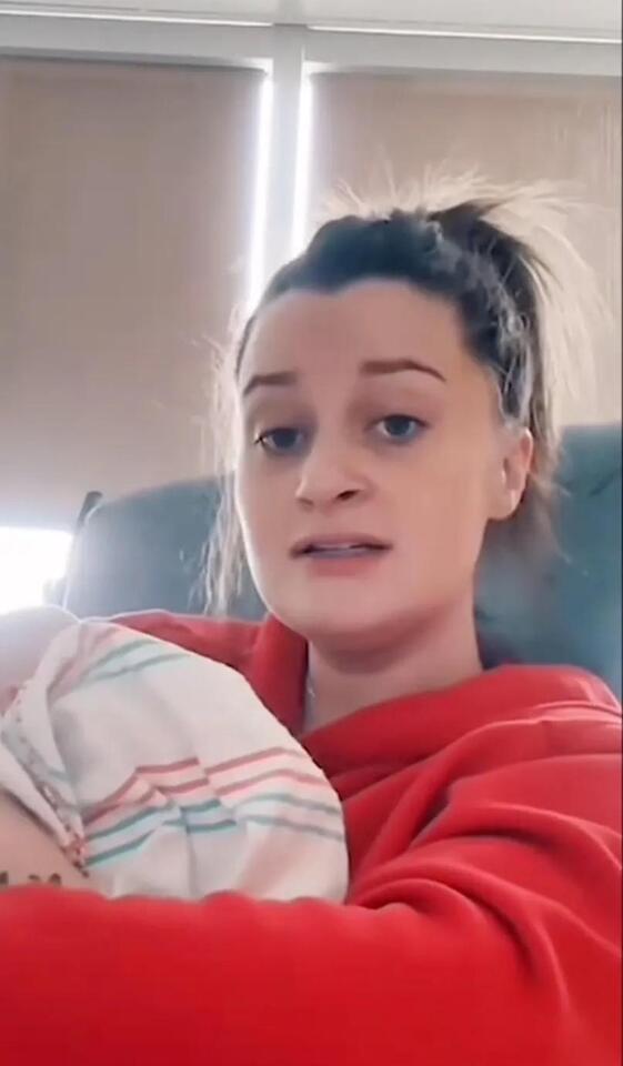 Alexandra Linsey Hill, in a red hoodie, holds her newborn wrapped in a striped hospital blanket. She speaks directly to the camera with a tense or defensive expression, likely during a livestream.