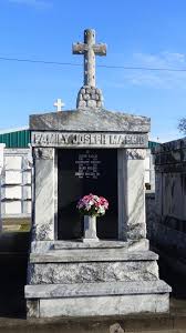 A marble tomb inscribed with "FAMILY JOSEPH MAGGIO" at the top, located in a New Orleans cemetery. The tomb has a large cross on top and a vase of pink and white flowers placed in the center beneath the family name.