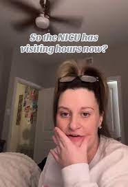 Alexandra Linsey Hill is indoors wearing a white sweater and glasses perched on her head. She rests her chin on her hand with a slight smile. The image includes overlaid text reading, "So the NICU has visiting hours now?" implying sarcasm.