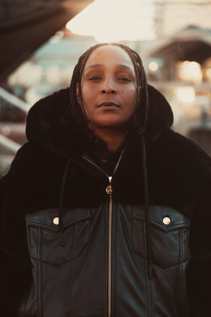 Felicia Pearson standing outdoors in a black fur-lined leather jacket with braids, looking directly into the camera with a calm, serious expression. The background is softly lit with warm city lights.
