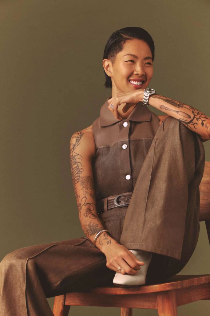 Kristen Kish sitting on a wooden chair with one leg up, dressed in a brown sleeveless button-up outfit, smiling with visible forearm tattoos and a silver watch.