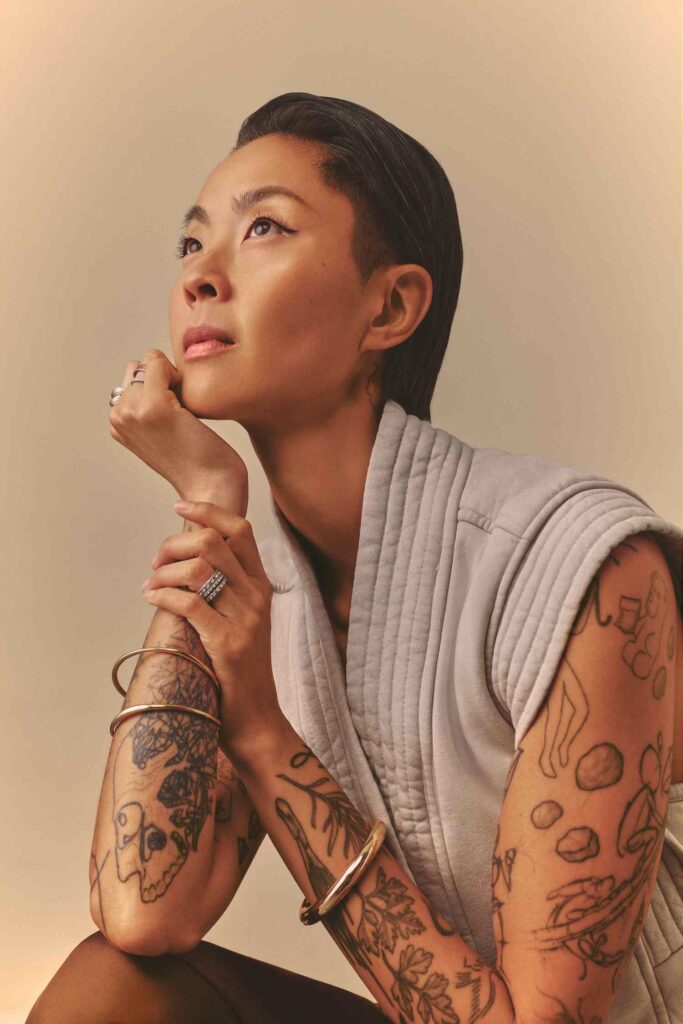 A thoughtful portrait of Kristen Kish looking upward, wearing a light gray textured top, resting her chin on her hand with tattoos and bracelets visible.