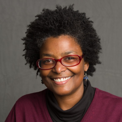 Mary L. Washington in a studio portrait setting, smiling, wearing red glasses and a burgundy top over a black cowl.