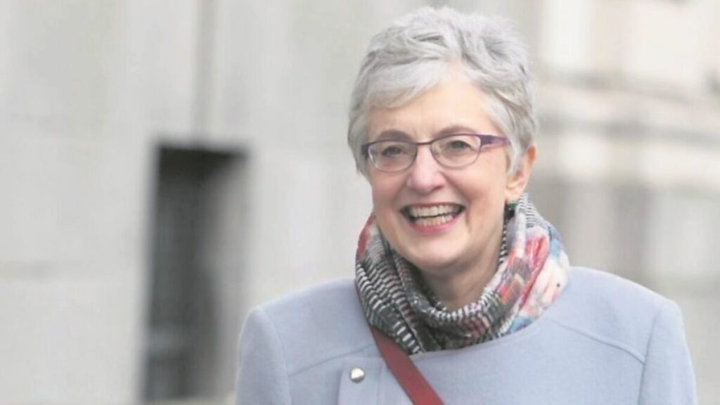 Katherine Zappone walking outside in a grey jacket and multicolored scarf, smiling in front of a stone building.