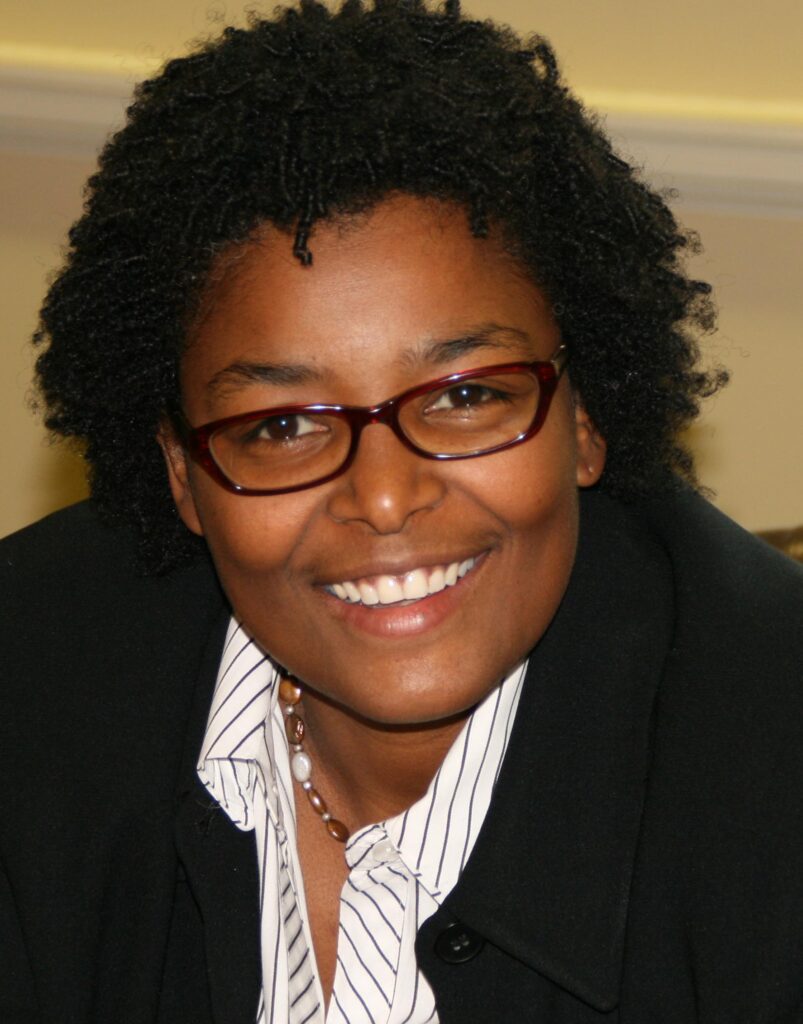 Mary L. Washington smiling, wearing a black jacket over a striped shirt with a pearl necklace and dark red glasses.