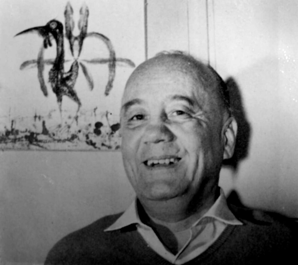 Black and white photo of Daniel Guérin smiling in front of an abstract painting featuring a bird-like creature.