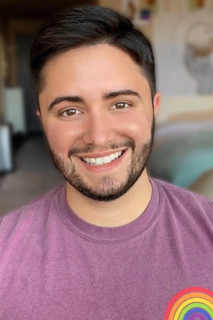Aiden Thomas smiles warmly while wearing a mauve shirt with a rainbow patch over the chest, standing in a softly lit room with blurred art and furniture in the background.