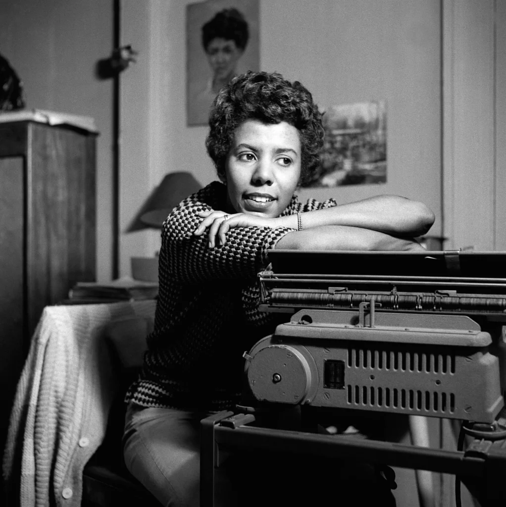 Lorraine Hansberry leans on a typewriter, smiling slightly, seated in a cozy room with framed artwork behind her. She wears a checkered top.