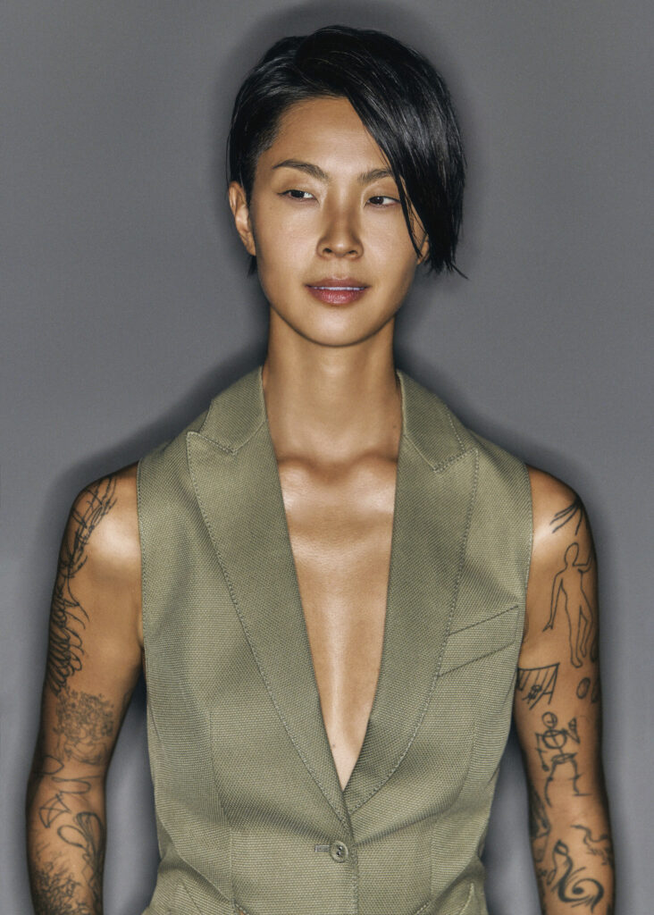 Kristen Kish in a muted green sleeveless tailored vest, standing against a gray backdrop with sleek side-parted hair and bold black ink tattoos on both arms.