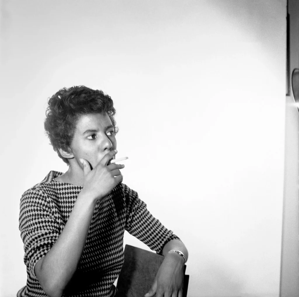 Lorraine Hansberry, wearing a checkered top, sits in front of a white backdrop, holding a cigarette and looking contemplatively to the side.