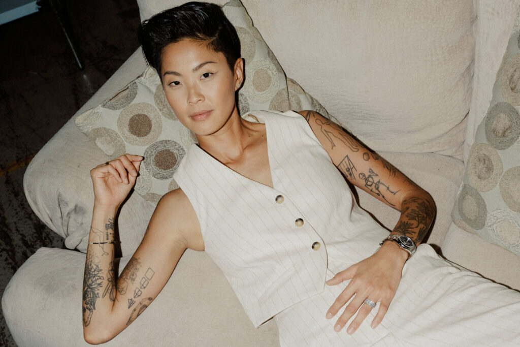 Kristen Kish reclining on a beige couch wearing a sleeveless cream pinstriped vest and pants, showcasing her tattooed arms and a wedding ring.