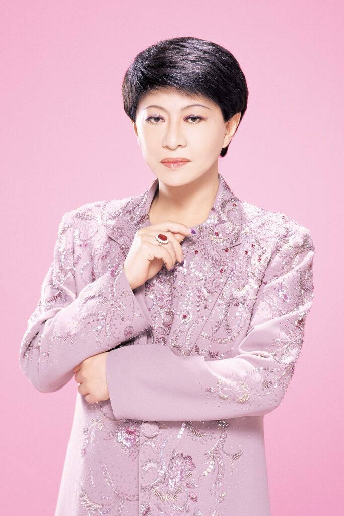 Kenichi Mikawa stands against a bright pink backdrop, dressed in a lavender jacket covered in glittering sequins and embroidery. His makeup is immaculate, and he holds one hand delicately over the other, showcasing a jeweled ring.