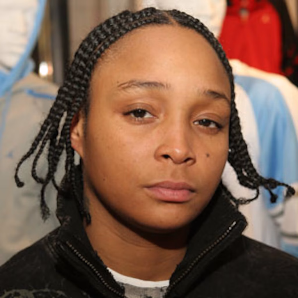 Close-up image of Felicia Pearson with a neutral expression, wearing a black zip-up jacket and white undershirt. Her hair is braided and she is looking slightly past the camera. The background is softly blurred, drawing focus to her face.