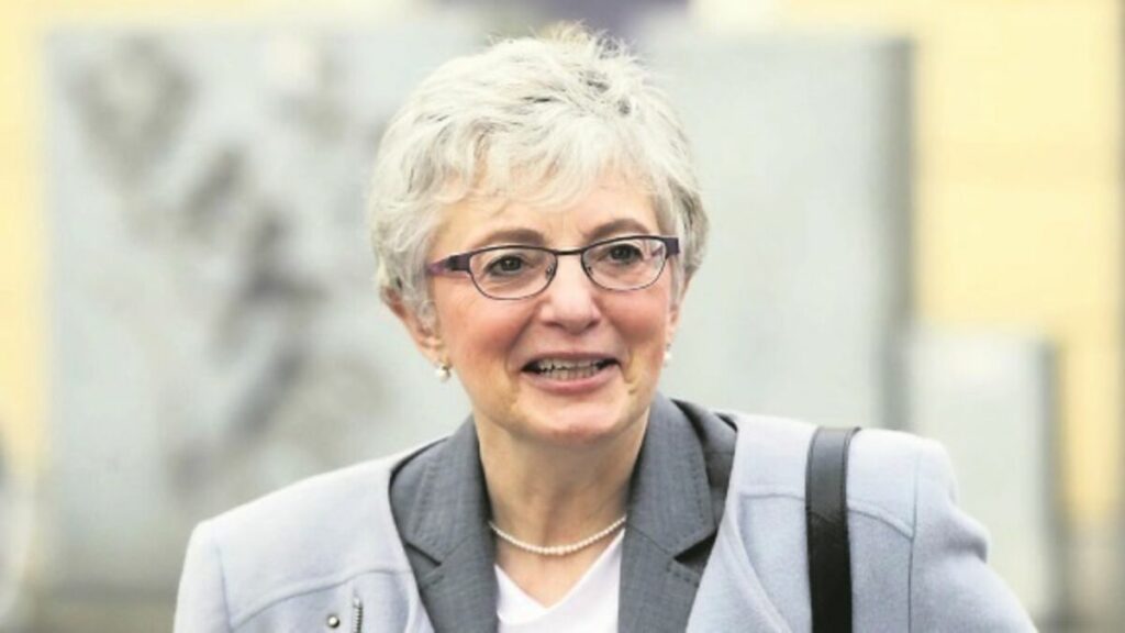Katherine Zappone outdoors wearing a light grey jacket, smiling while speaking, with buildings blurred in the background.